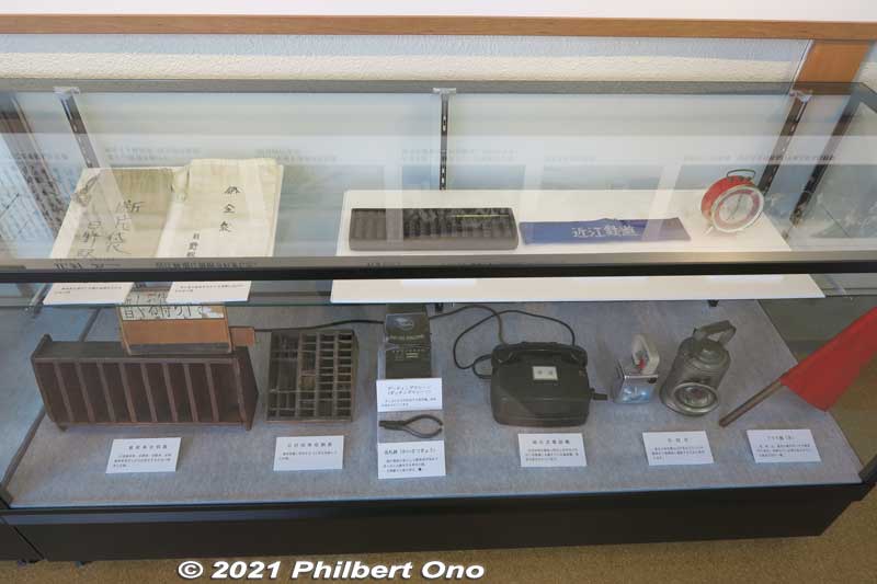 Hino Station Railway Museum exhibits. Things used by Hino Station staff. Lower shelf has a train ticket holder, rubber date stamp holder, ticket dating machine, ticket clippers, magneto telephone with a crank, signal light, and red signal flag.
